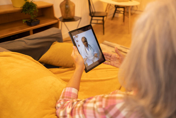 Senior woman seated on a couch, having a telehealth consultation with a doctor via a tablet. - Photo, Image