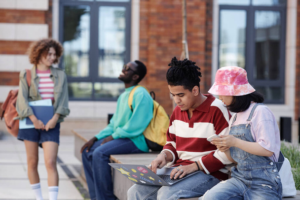 Monipuolinen ryhmä nuoria opiskelijoita istuu rivissä ulkona college kampuksella ja opiskelee kannettava tietokone, kopioi tilaa - Valokuva, kuva