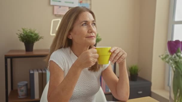 Kobieta w średnim wieku pije filiżankę kawy w domu, przywołując przytulną i relaksującą atmosferę wewnątrz. - Materiał filmowy, wideo