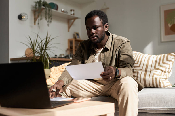 Portrait d'un homme noir adulte utilisant un ordinateur portable tout en travaillant de la maison dans la lumière du soleil et en tenant un document, un espace de copie - Photo, image