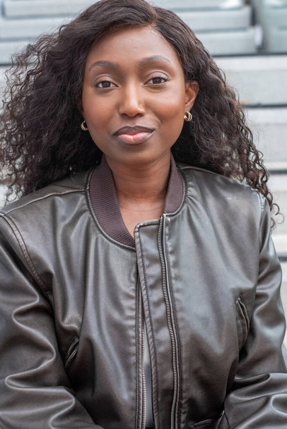 This image features a Black woman posing outdoors, wearing a stylish leather jacket. Her curly hair cascades around her face, and she gazes confidently at the camera. She accessorizes with gold hoop - Photo, Image