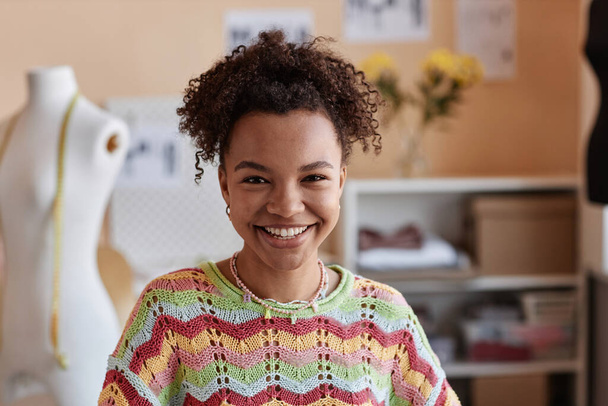 Junge lächelnde Modedesignerin im Strickpullover blickt in Werkstatt gegen Schaufensterpuppe und Nähbedarf in die Kamera - Foto, Bild
