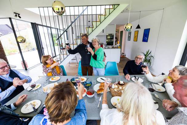 Група літніх людей, які збираються, насолоджуючись їжею та вином у сучасній їдальні, наповненій сміхом та веселими - Фото, зображення