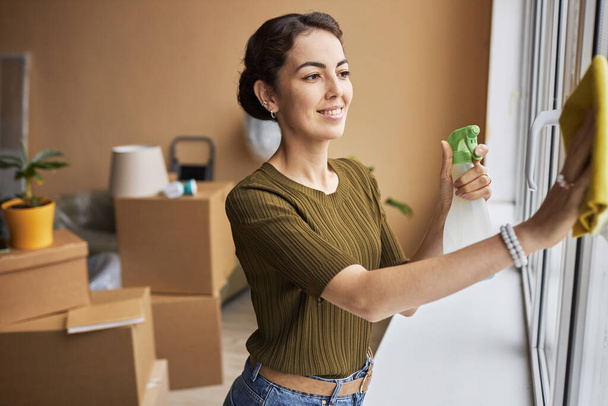 Jovem mulher sorridente com detergente spray limpando janelas com espanador enquanto está em pé na espaçosa sala de estar do novo apartamento - Foto, Imagem