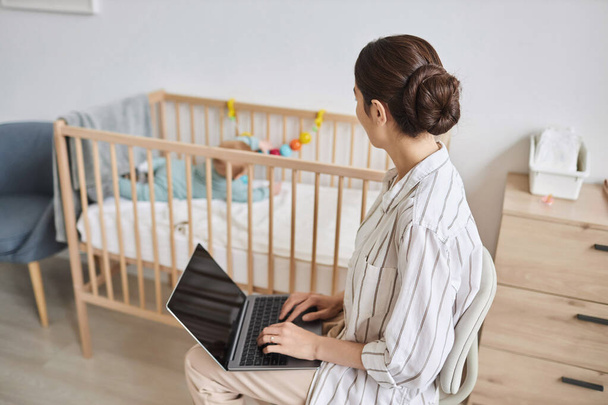 Side view portrait of young mother using laptop at home or doing internet research on parenting and watching baby, copy space - Photo, Image