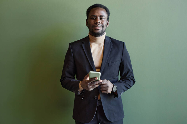Mittlere Aufnahme eines lächelnden Afroamerikaners im modischen Businessanzug, der sein Smartphone in die Kamera hält, die vor einer grünen Wand steht - Foto, Bild