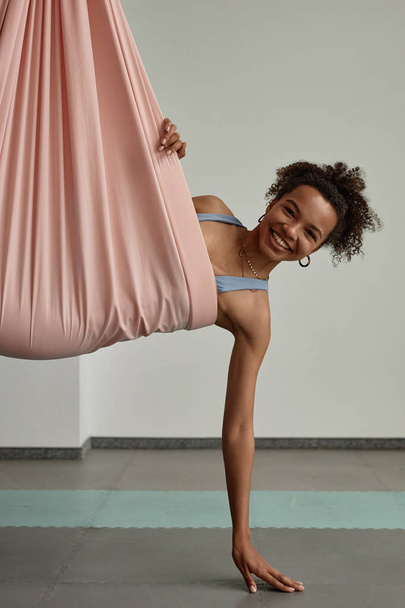 Ritratto verticale di giovane donna nera che si gode lo yoga aereo in amaca rosa e sorride alla macchina fotografica - Foto, immagini