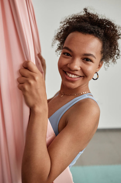Portrait de jeune femme noire souriant à la caméra dans un hamac rose profitant d'un cours de yoga aérien - Photo, image