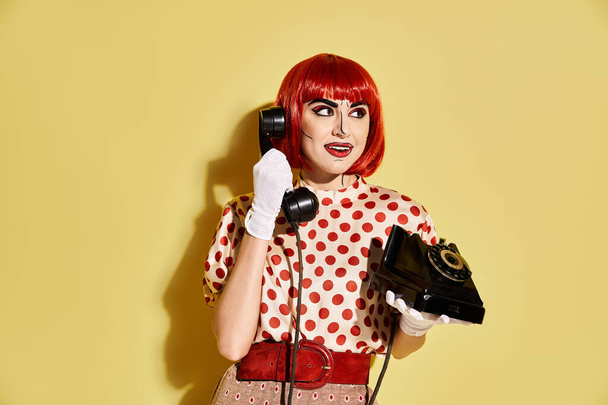 Una pelirroja vibrante con maquillaje pop art y blusa de lunares sostiene un teléfono sobre un atrevido fondo amarillo. - Foto, imagen