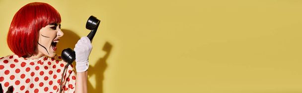 Uma mulher ruiva criativa com maquiagem pop art mantém o telefone de forma lúdica em um fundo amarelo brilhante. - Foto, Imagem
