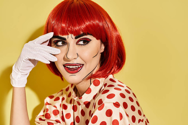 Een mooie roodharige vrouw in een polka dot shirt en witte handschoenen, met creatieve pop art make-up, staat tegen een gele achtergrond. - Foto, afbeelding