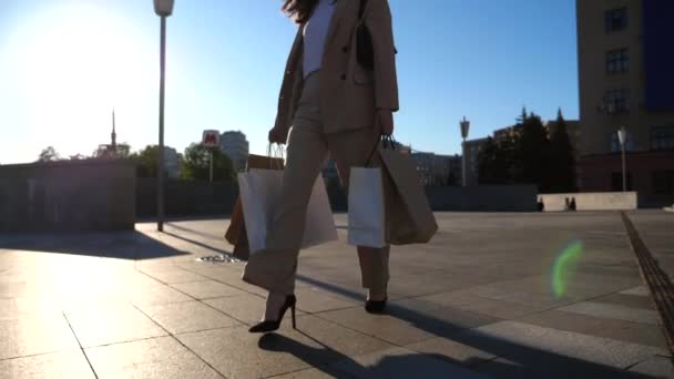 Elegant young lady holds shopping bags going at city square after purchases. Attractive woman carries paper packets walking along sunny urban street. Sales and discounts concept. Slow motion. - Video, Çekim