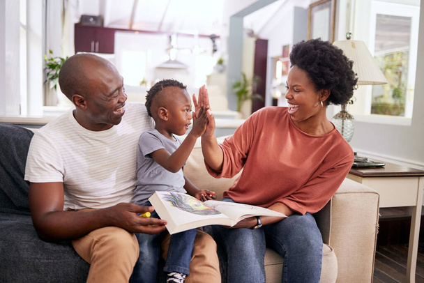 Black family, high five and happy in living room with child, color book and motivation for kid. Lounge, woman and man with smile, together or parents with boy, excited and drawing of youth on couch. - Fotografie, Obrázek