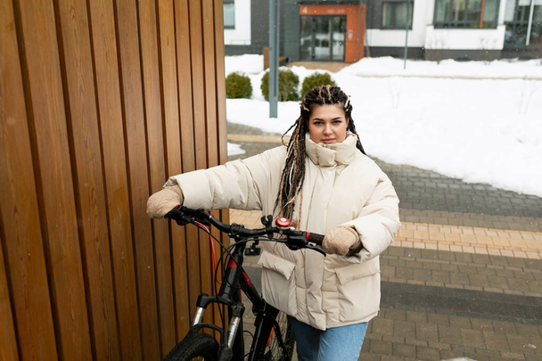 Une femme avec des dreadlocks est debout à côté d'un vélo. Elle a l'air de regarder quelque chose hors caméra, les mains posées sur le guidon. Le fond montre un cadre de rue avec - Photo, image