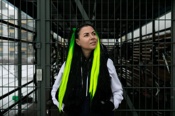 A woman with bright green hair stands confidently in front of a rustic wooden fence, her striking appearance contrasting with the natural surroundings. She gazes directly at the viewer, exuding a - Photo, Image