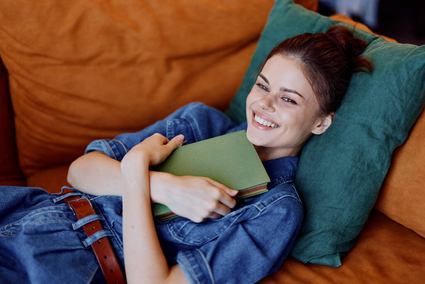 Ευτυχισμένη γυναίκα ξαπλωμένη στον καναπέ με βιβλίο και μεγάλο χαμόγελο στο πρόσωπό της, απολαμβάνοντας μια χαλαρωτική στιγμή στο σπίτι - Φωτογραφία, εικόνα