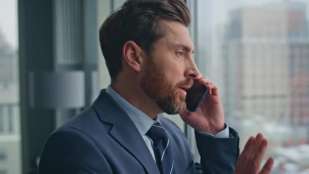 Annoyed boss calling cellphone gesturing hand in cityscape office closeup. Angry bearded businessman feeling stress at mobile phone conversation standing at big windows. Emotional entrepreneur call  - Footage, Video