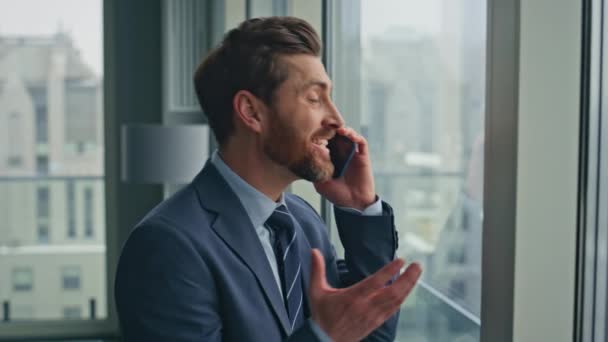 Успішний інвестор, який говорить смартфон, виглядає великим вікном крупним планом. Щасливий бородатий підприємець чує, як хороші новини жестикулюють по телефону. Усміхнений впевнений бізнесмен дзвонить в розкішний офіс  - Кадри, відео