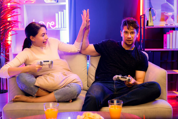 Ζευγάρι χαρούμενη του παίκτη βιντεοπαιχνίδι στην τηλεόραση χρησιμοποιώντας joysticks κάνοντας κόλλα πέντε του νικητή μαζί στο παιχνίδι ανταγωνισμού στο στούντιο δωμάτιο σε κόκκινο μπλε λάμπα νέον στο άνετο σπίτι. Αξίωμα. - Φωτογραφία, εικόνα