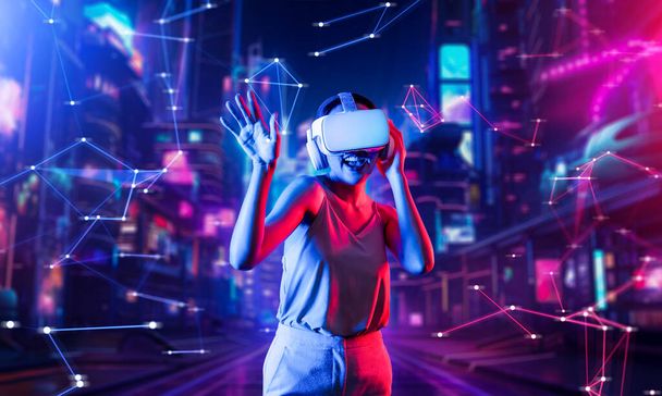 Kobieta stojąca w wirtualnej rzeczywistości w cyberpunkowym budynku nosi zestaw słuchawkowy VR łączący metawersy, przyszłą technologię społeczności cyberprzestrzeni, kobieta tańcząca trzymając gogle za jedną rękę. omamy. - Zdjęcie, obraz