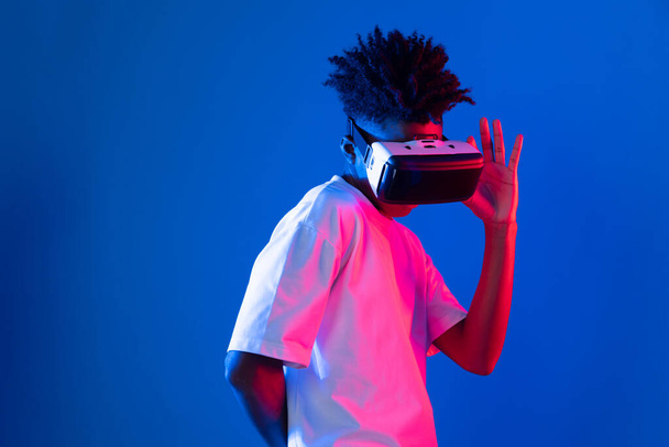 Удивлённый молодой афроамериканец, смотрящий через виртуальную реальность с интересной объектной голограммой на розовой синей неоновой стене в метавселенной, соединяющей цифровую футуристическую технологию с виртуальной реальностью. Вклад. - Фото, изображение