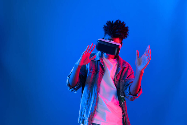 Lachende jonge Afro-Amerikaanse kijkend door VR dansen in lied met hologram gaming op roze blauwe neon muur op metaverse wereld verbinden digitale futuristische technologie virtual reality. Vooruitzichten. - Foto, afbeelding