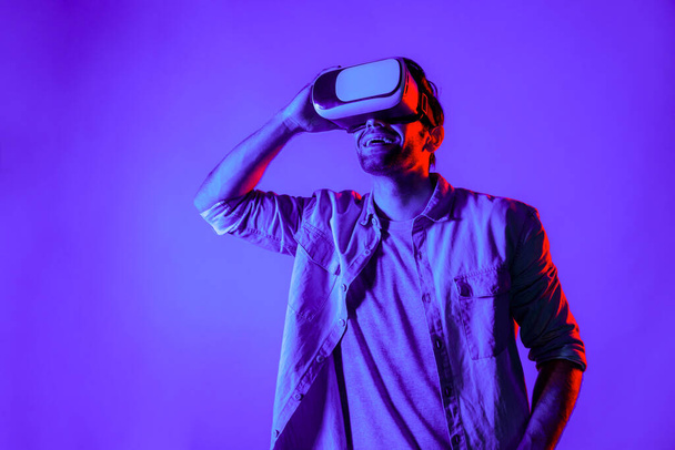 Man with VR goggles and looking in metaverse or simulated virtual world πρόγραμμα σε φόντο νέον φωτός. Καυκάσιος έφηβος αγγίζει τα ακουστικά εικονικής πραγματικότητας ενώ χρησιμοποιεί τεχνολογική καινοτομία. Απόκλιση. - Φωτογραφία, εικόνα