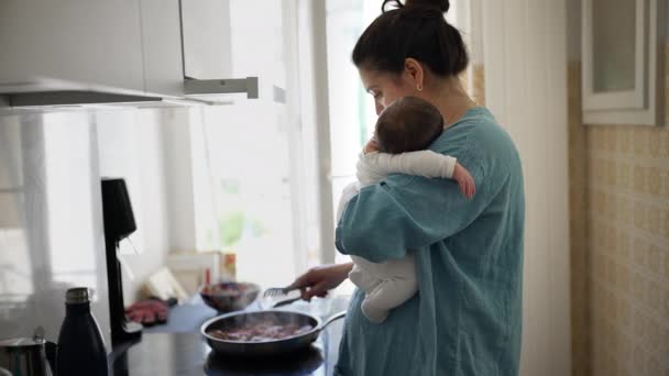 Mama w niebieskiej koszuli trzymająca dziecko podczas mieszania jedzenia na patelni na kuchence, oświetlona naturalnym światłem przez okno, pokazując wielozadaniowość, opiekę i równoważenie rodzicielstwa z codziennymi obowiązkami - Materiał filmowy, wideo