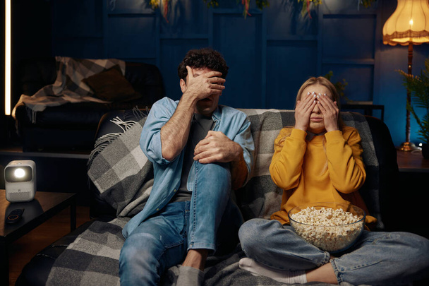 Man and woman are sitting on a couch covering their eyes with their hands. Couple watching scary movie together - Фото, изображение