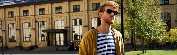Egy divatos fiatalember, vörös hajjal, aki a városban sétál, sárga dzsekiben és napszemüvegben.. - Fotó, kép