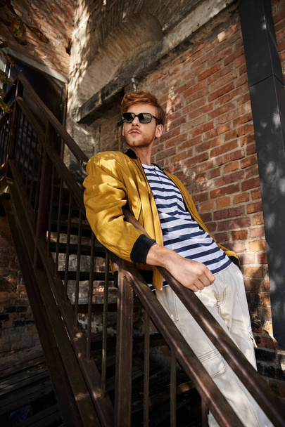 Stylish red-haired man in yellow jacket leans on railing in city. - Photo, Image