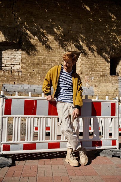 Joven hombre pelirrojo vestido con un atuendo rubio sentado con confianza sobre una cerca blanca en la ciudad. - Foto, imagen