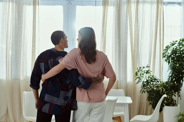 Una pareja gay con atuendo casual se para lado a lado, mirando por una gran ventana en una acogedora casa. - Foto, imagen
