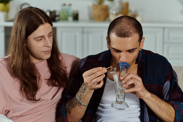 Una coppia gay in abbigliamento casual rilassarsi su un divano a casa, uomo illuminazione marijuana nel bong di vetro - Foto, immagini