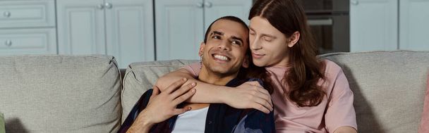 Ein schwules Paar in lässiger Kleidung umarmt sich auf einer bequemen Couch zu Hause und teilt einen Moment der Liebe und Zuneigung. - Foto, Bild