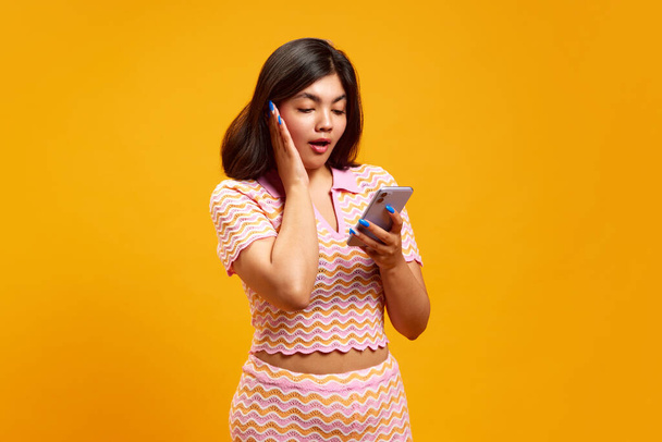 Mujer india joven sosteniendo teléfono inteligente y mirando a su pantalla con expresión conmocionada contra el fondo vibrante estudio amarillo. Concepto de emociones humanas, moda y estilo, belleza, internet. - Foto, Imagen