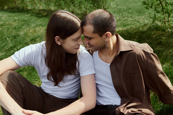 A gay couple in casual clothes enjoys a peaceful moment together, sitting on the grass in nature. - Фото, изображение