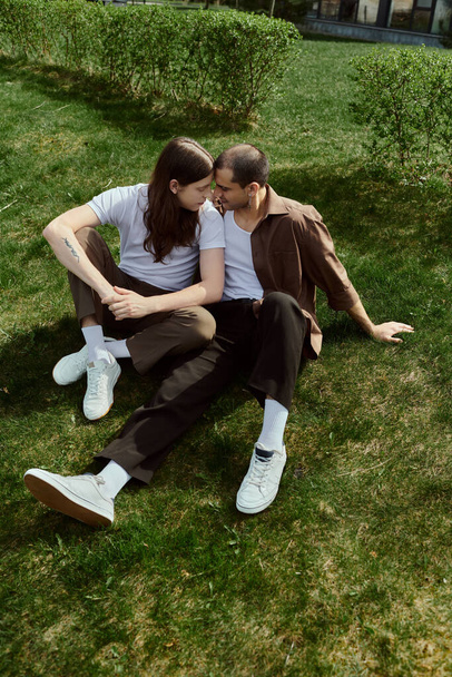 Ein schwules Paar, lässig gekleidet, genießt einen ruhigen Moment zusammen, während sie auf dem Gras sitzen. - Foto, Bild