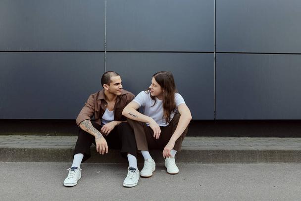 Ein schwules Paar in legerer Kleidung sitzt friedlich nebeneinander auf dem Boden im Freien. - Foto, Bild