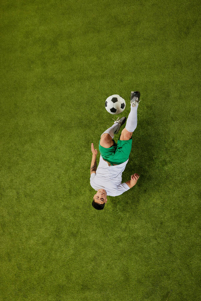 Aerial shot of soccer player in mid-air performing acrobatic kick, with ball suspended above green field. Concept of professionals sport, competition, tournament, energy, action. Ad - 写真・画像