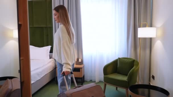 Gyönyörű női tripper divatos ruhában belép egy elegáns szállodai szobába, begöngyölíti a bőröndjét, és elkezdi kicsomagolni. A természetes fény nagy ablakokon keresztül tölti meg a teret.. - Felvétel, videó