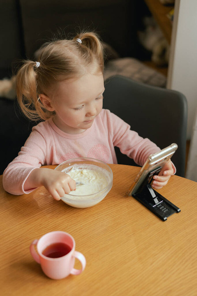 Baby girl eats breakfast and looks at the phone at the table. Harmful upbringing of a child with gadgets. Children and technology or Healthy food for kid concept. Part of the series. - Photo, Image
