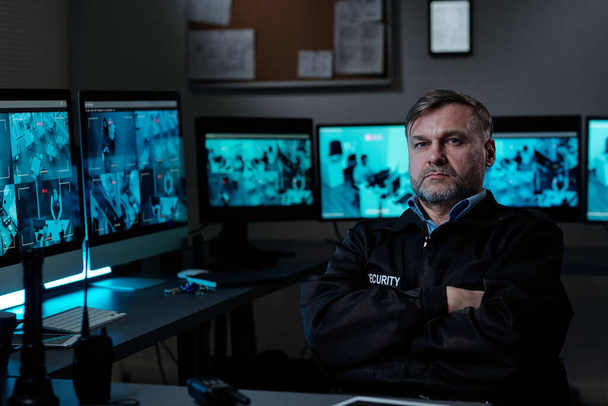 Серйозний зрілий охоронець тримає руки схрещені грудьми, сидячи на робочому місці з ліхтарем, Walkie-talkie і комп'ютерами - Фото, зображення