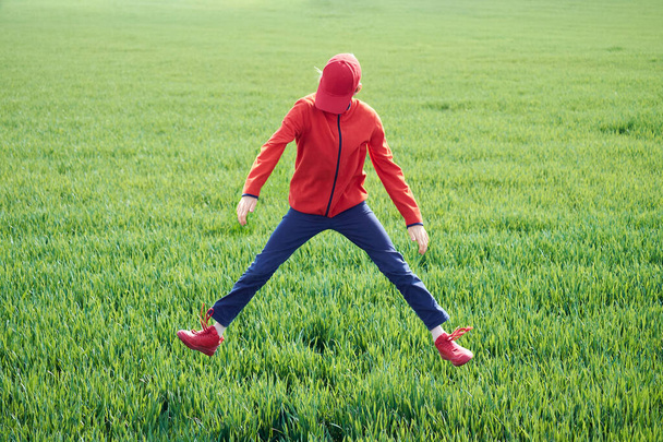 Nastolatek skacze z rozłożonymi nogami na tle zielonego pola trawy. - Zdjęcie, obraz