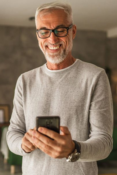 Jeden dojrzały mężczyzna kaukaski w okularach trzymać telefon komórkowy i odkrywać internet lub SMS w domu - Zdjęcie, obraz