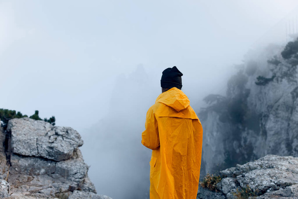 Parlak sarı yağmurluklu yalnız bir figür gizemli sisle örtülü görkemli dağ zirvesini fethediyor. - Fotoğraf, Görsel