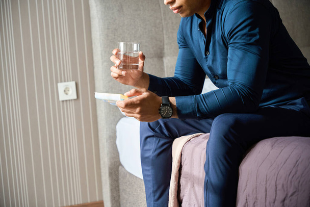 Cropped image of african american businessman with glass of water and pills sitting on bed in hotel room at daytime. Concept of health care - Photo, Image