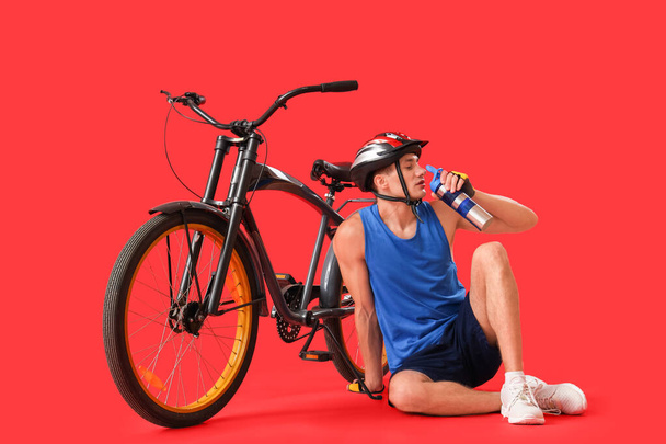 Young man sitting near bicycle and drinking water on red background - Photo, Image