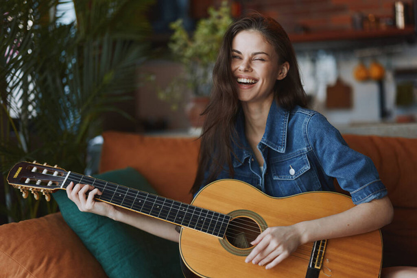Donna felice seduta su un divano a suonare la chitarra acustica con un sorriso sul viso - Foto, immagini