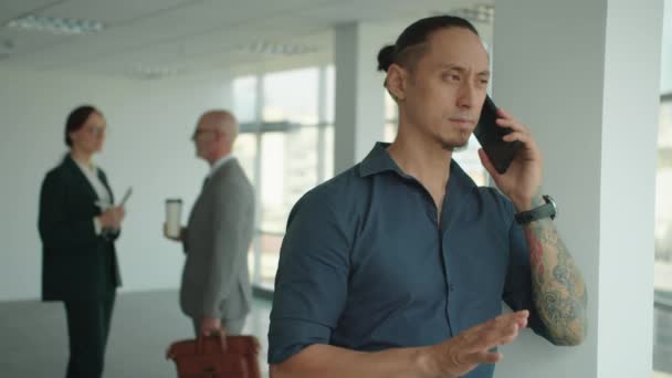 Multiethnisch tätowierte männliche Unternehmer ruft Kunden am Handy an, wenn sie an einem modernen Arbeitsplatz arbeiten - Filmmaterial, Video